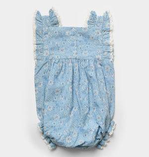 The Early Sunday Baby Girls’ Eva Baby Romper features a blue floral print with lace trim, ruffled shoulder details, and button closures on the back, shown against a white background. This essential piece combines charming style and everyday comfort for your little one.