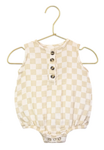 The Lou Lou & Company Baby Cloud Muslin Bubble Romper features a beige and white checkered sleeveless design with four front buttons, elastic leg openings, and convenient bottom snaps for easy changes. Perfect for warm weather, this essential romper is displayed on a gold hanger against a white background.