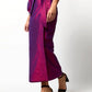 A person stands against a plain white background wearing the Ann Mashburn Kira Silk Shantung Wrap Skirt in shiny magenta with an adjustable tie waist, a matching puff-sleeve top, and black open-toe heeled sandals.