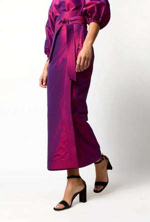 A person stands against a plain white background wearing the Ann Mashburn Kira Silk Shantung Wrap Skirt in shiny magenta with an adjustable tie waist, a matching puff-sleeve top, and black open-toe heeled sandals.