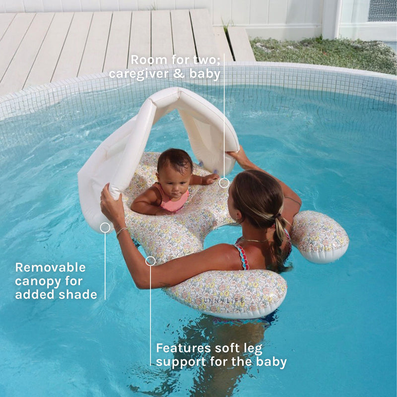 A parent and baby enjoy the Sunnylife Garden Ditsy Floral Float Together Baby Seat—a dual float designed for two, featuring a charming flower pattern, removable shade canopy, and supportive leg holes for baby—in a clear, round pool. Bold text emphasizes the float’s roomy design for adult and child, the easy-to-remove shade canopy, and specially engineered support for baby’s legs.