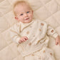 A baby lies on a cream-colored quilt, dressed in the Quincy Mae Baby Boys’ and Boys’ Relaxed Fleece Sweatshirt—a cozy essential featuring playful bear faces and animal prints—looking up with a slight smile.