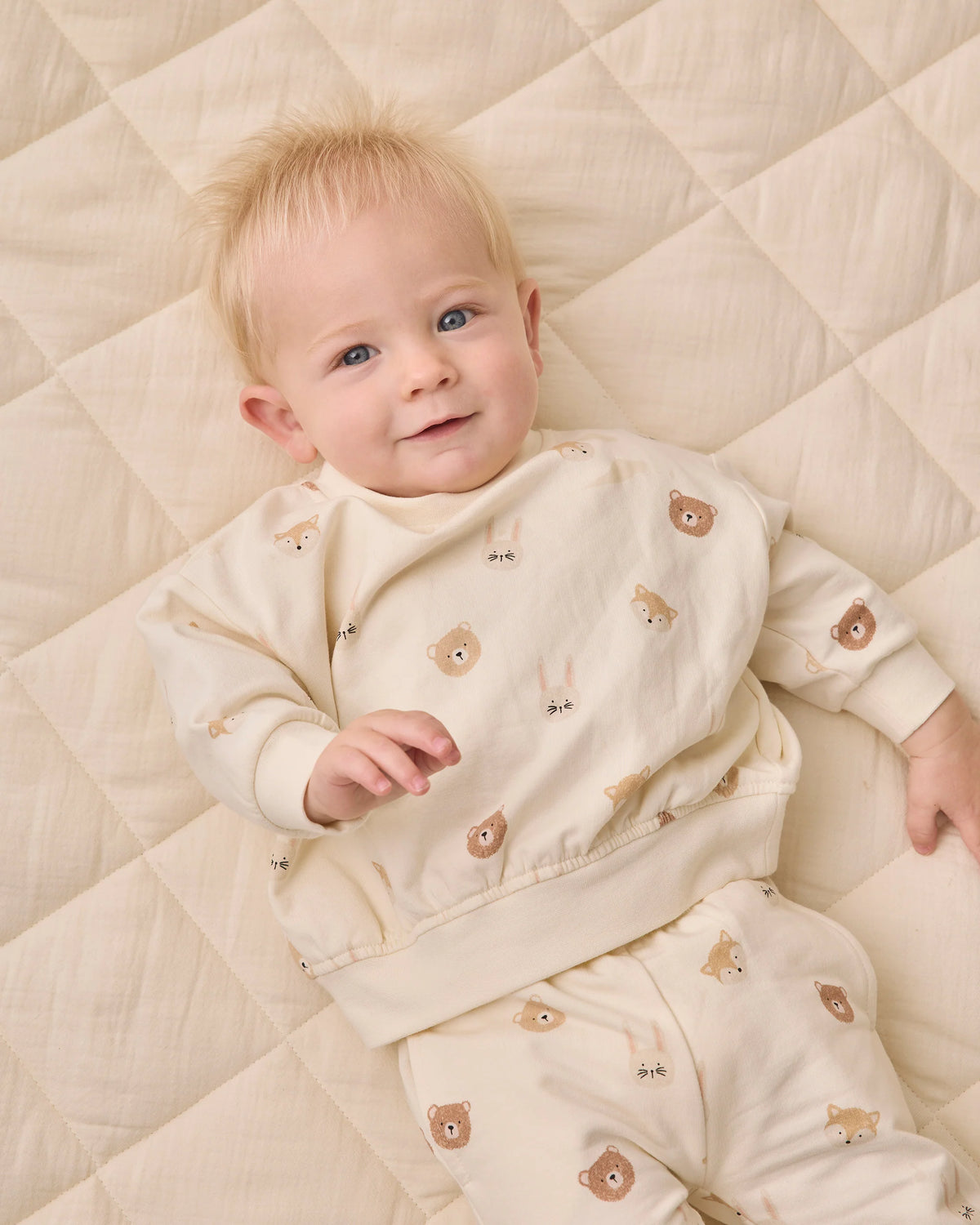 A baby lies on a cream-colored quilt, dressed in the Quincy Mae Baby Boys’ and Boys’ Relaxed Fleece Sweatshirt—a cozy essential featuring playful bear faces and animal prints—looking up with a slight smile.