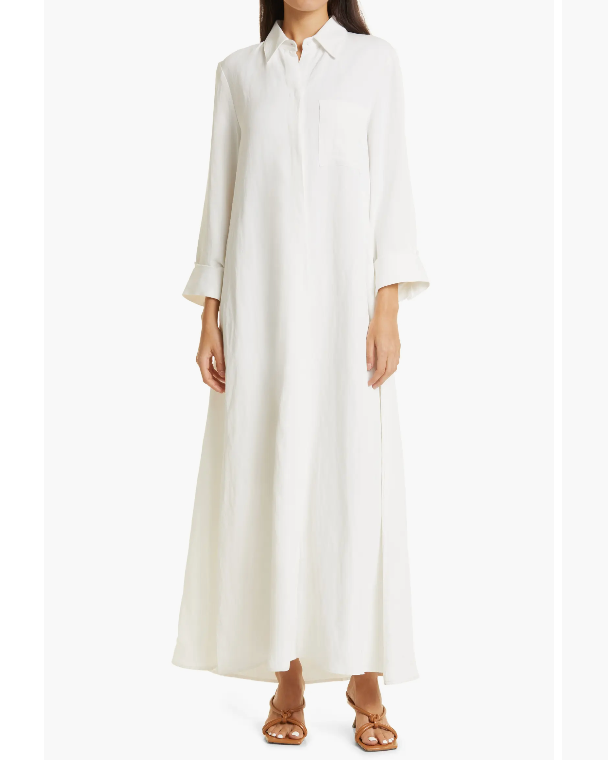 A person wears Jenny's Coated Linen Gown by TWP, an oversized, A-line shirt dress in long, white, button-up viscose linen, paired with brown sandals.