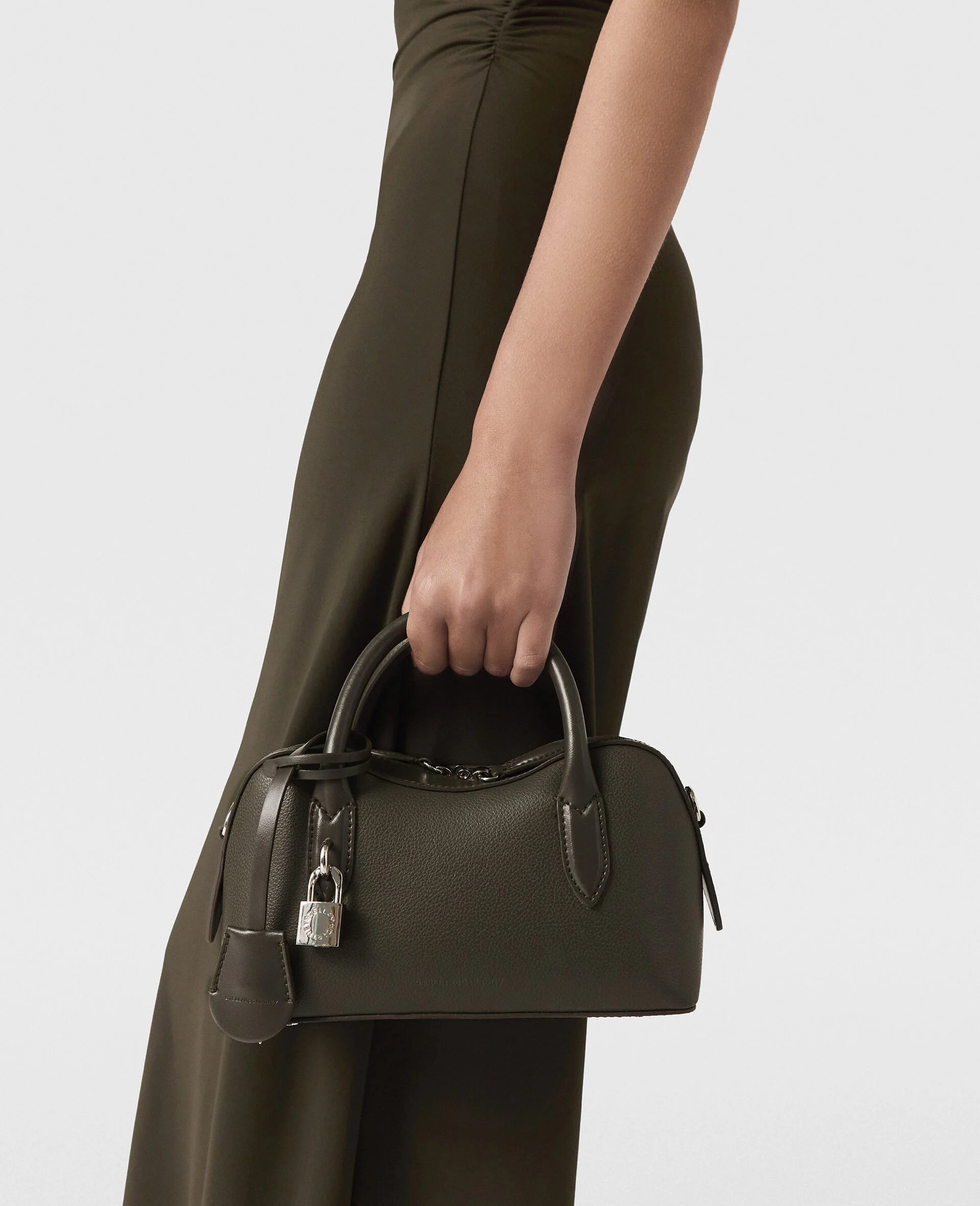A person in an olive dress holds the Stella McCartney Ryder Crossbody Bag, a small structured Italian-crafted bag with lock and key charm, set against a plain light background.