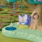The Calico Critters Woodland Wonder Fairy Castle features a small fairy figurine with wings sitting beside a blue pond and a tree adorned with crystal decorations, creating a charming fairy forest scene. A pink animal figure is nestled in the tree hollow, adding to the enchanting collection of fairy accessories included in this magical set.