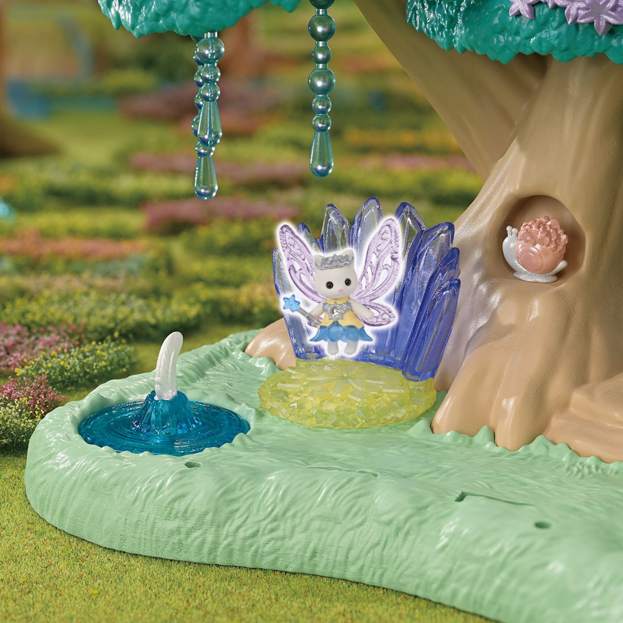 The Calico Critters Woodland Wonder Fairy Castle features a small fairy figurine with wings sitting beside a blue pond and a tree adorned with crystal decorations, creating a charming fairy forest scene. A pink animal figure is nestled in the tree hollow, adding to the enchanting collection of fairy accessories included in this magical set.