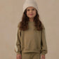 A young child wears the Rylee & Cru Baby Girls’ and Girls’ Oversized Sweatshirt in olive green with matching pants featuring white trim, paired with a cream knit beanie, standing against a plain beige background.