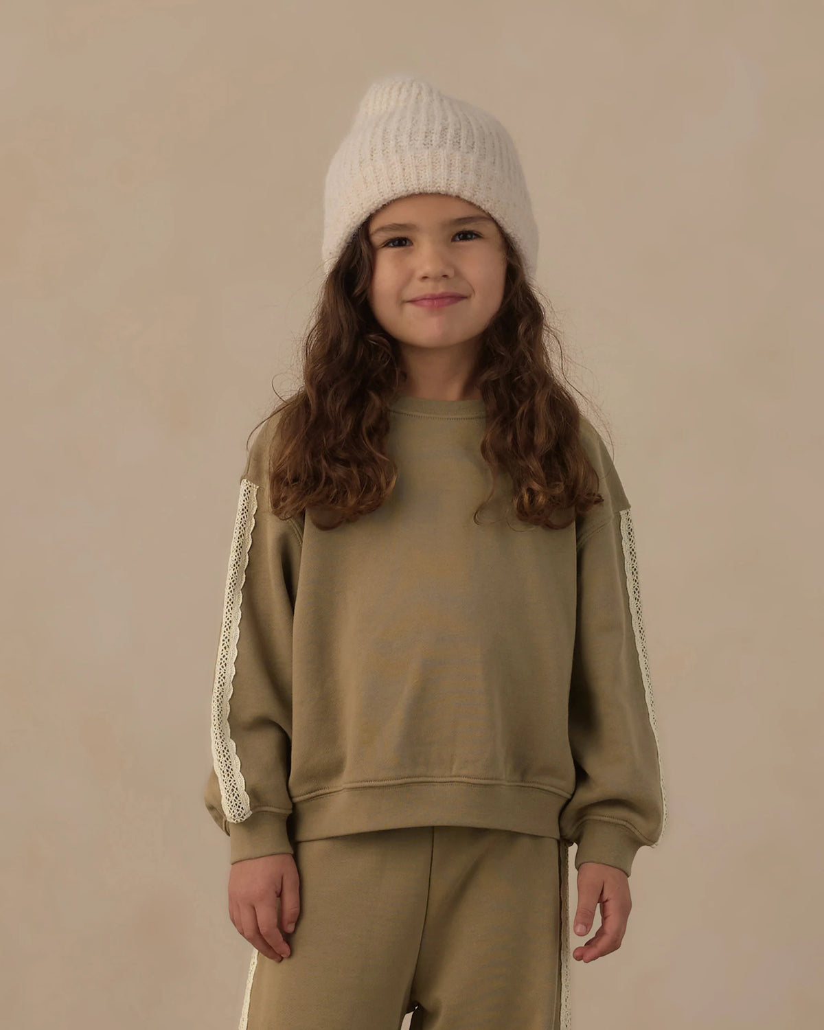 A young child wears the Rylee & Cru Baby Girls’ and Girls’ Oversized Sweatshirt in olive green with matching pants featuring white trim, paired with a cream knit beanie, standing against a plain beige background.