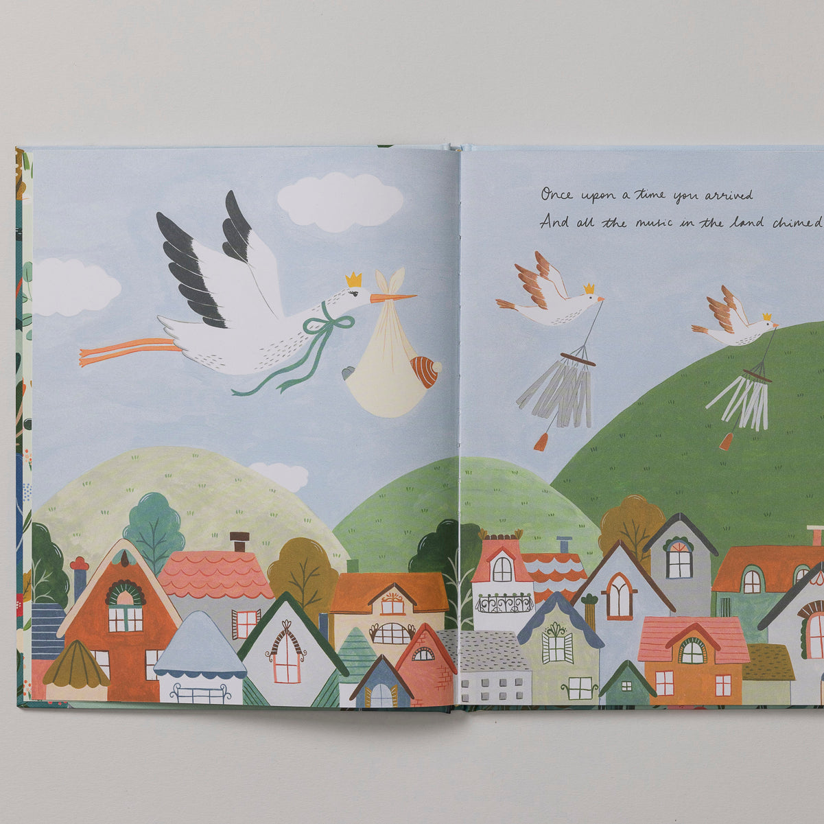 Pages from the charming children's book, *Write to Me Your Baby Story* by Write To Me, beautifully depict storks carrying bundles over a vibrant village. This delightful read is ideal for celebrating first birthdays. Storks glide above houses, hills, and trees while featuring an enchanting rhyming text.