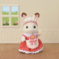 A Calico Critters bunny from the Village Cake Shop Starter Set, dressed in a red polka dot dress with a white apron and frilly hat, stands indoors on a brown floor with beige walls and a window in the background, looking ready to help at the cake shop or arrange charming decorations for sponge cakes.