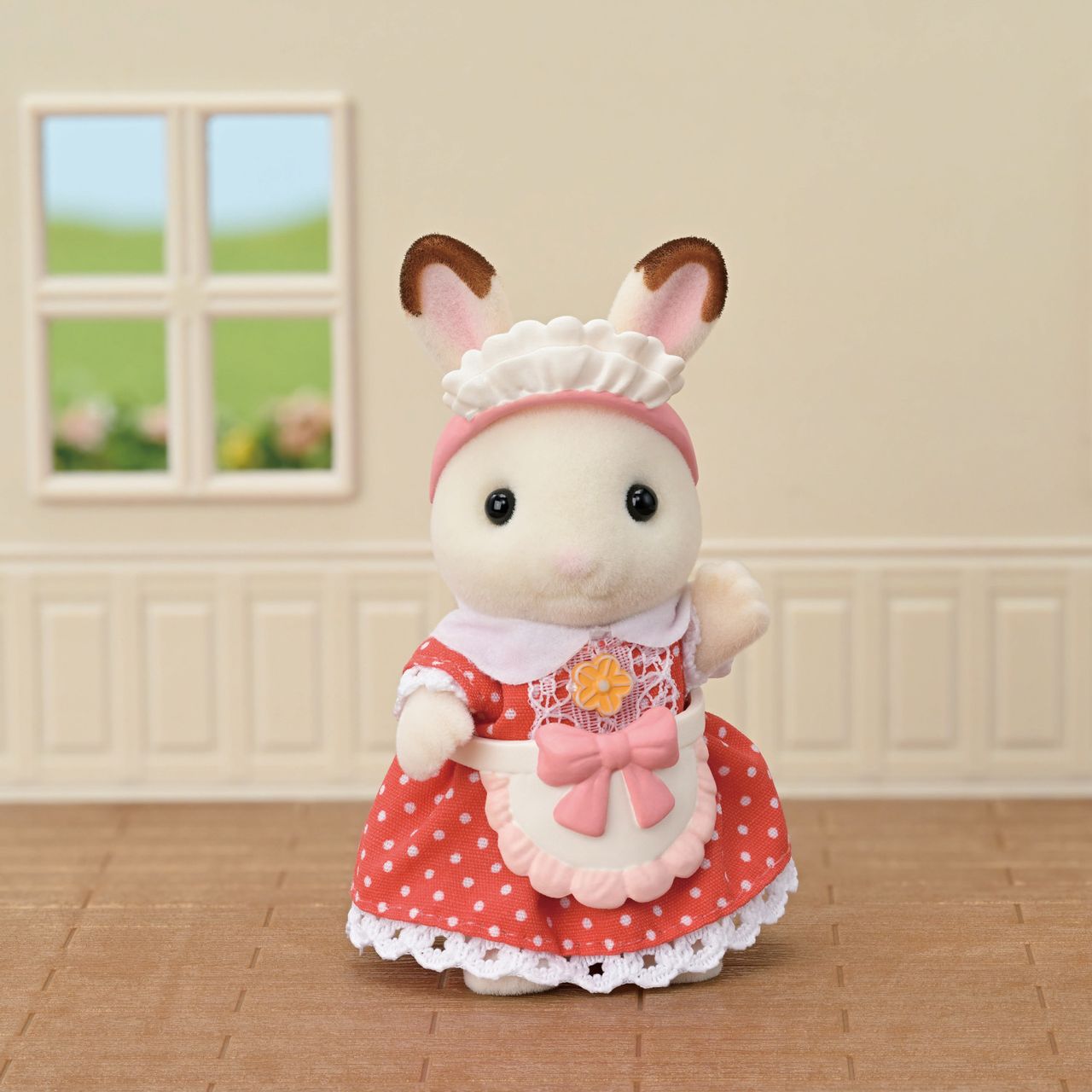 A Calico Critters bunny from the Village Cake Shop Starter Set, dressed in a red polka dot dress with a white apron and frilly hat, stands indoors on a brown floor with beige walls and a window in the background, looking ready to help at the cake shop or arrange charming decorations for sponge cakes.
