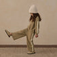 A young child wearing the Rylee & Cru Girls’ Wide Leg Sweat Pant in olive green, styled with a cozy oversized sweatshirt, beige knit hat, and coordinating slip-on shoes, stands playfully on a patterned rug with one leg lifted against a plain wall.