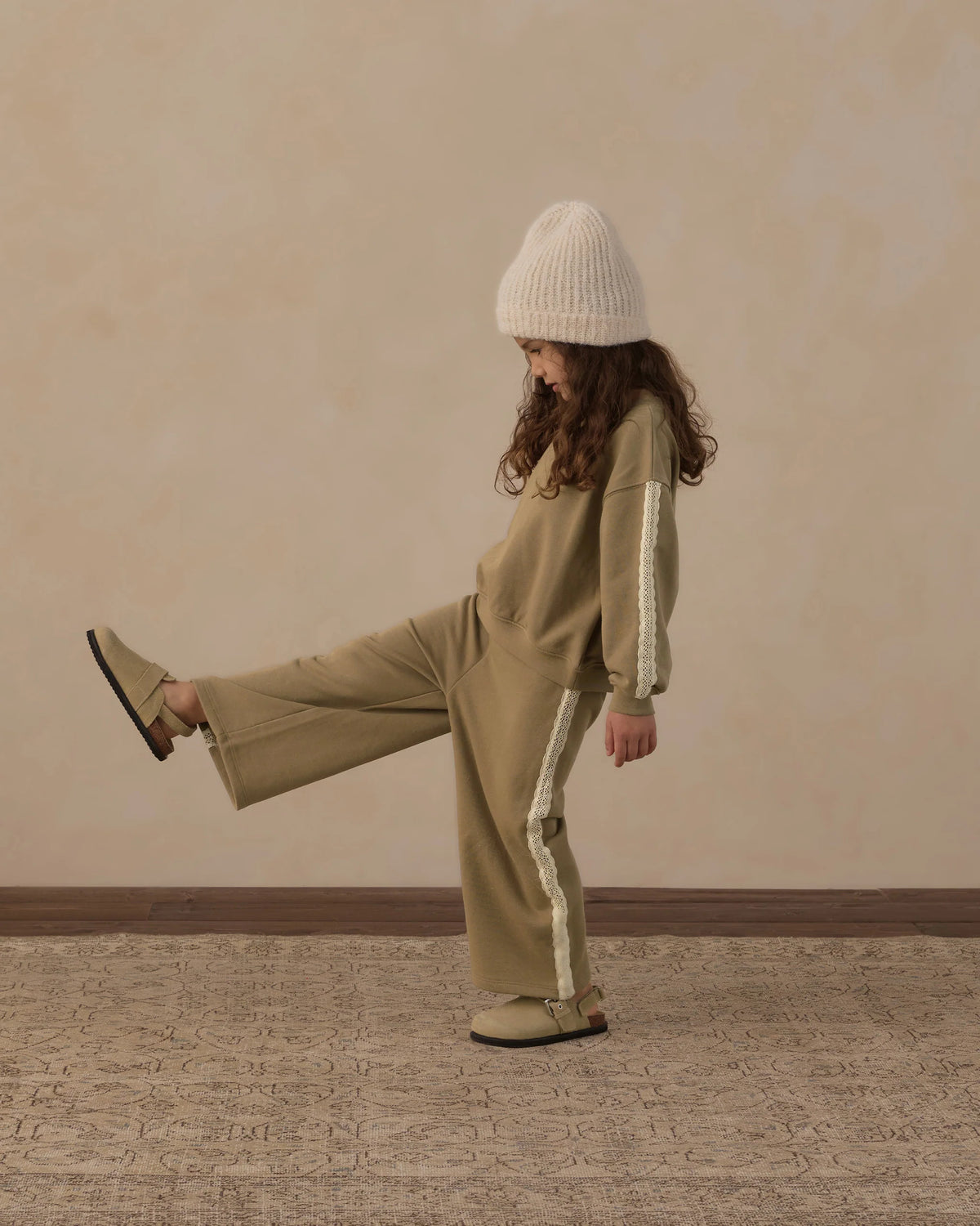 A young child wearing the Rylee & Cru Girls’ Wide Leg Sweat Pant in olive green, styled with a cozy oversized sweatshirt, beige knit hat, and coordinating slip-on shoes, stands playfully on a patterned rug with one leg lifted against a plain wall.