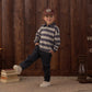 A child wears a striped shirt, a Rylee & Cru Baby Boys’ and Boys’ Kalen Pant featuring an adjustable fit, and a cap while standing indoors on a rug with one leg raised. Stacks of books and a lantern sit beside a wooden wall in the background.