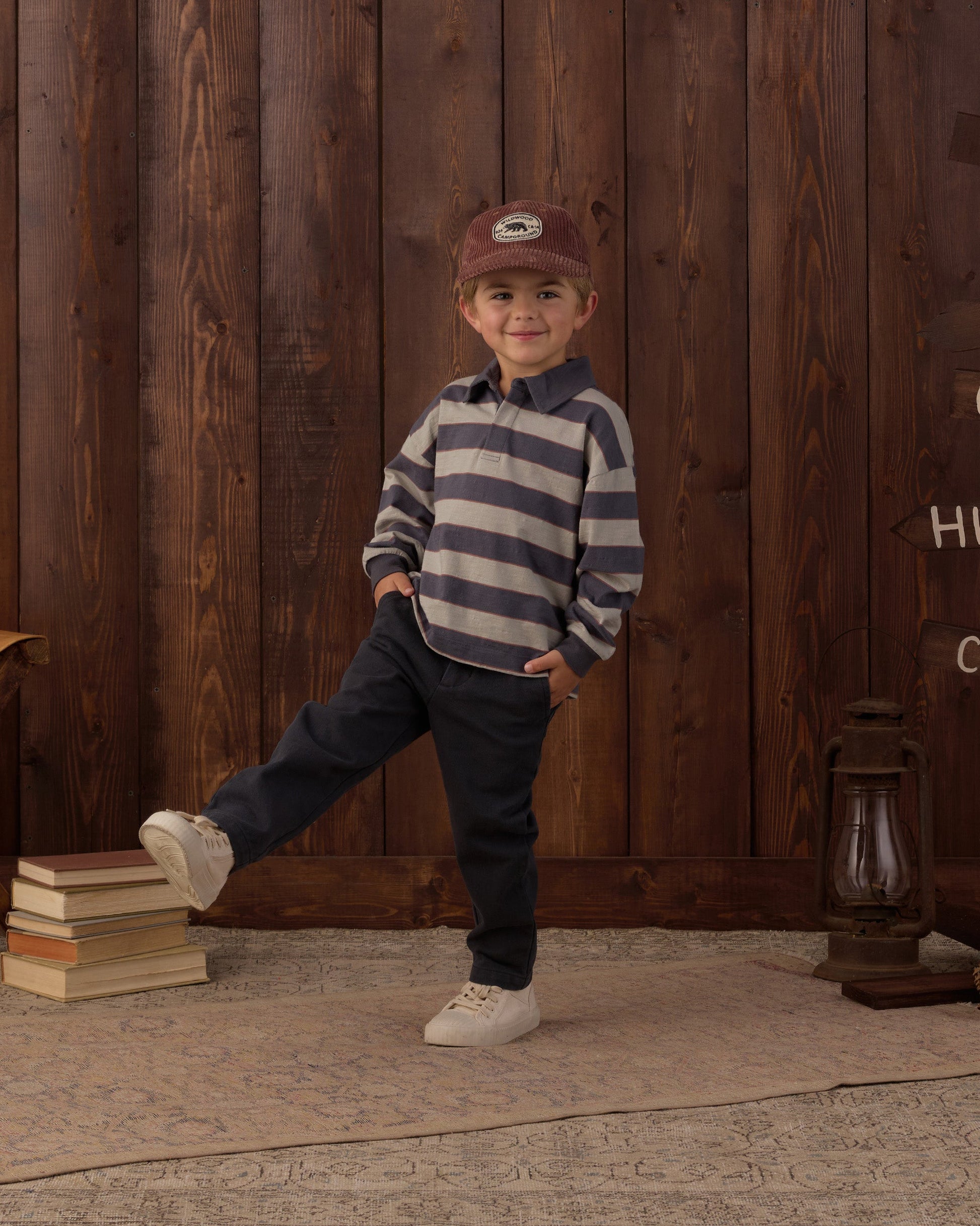 A child wears a striped shirt, a Rylee & Cru Baby Boys’ and Boys’ Kalen Pant featuring an adjustable fit, and a cap while standing indoors on a rug with one leg raised. Stacks of books and a lantern sit beside a wooden wall in the background.