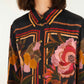 A woman wears the Farm Rio Chenille Flowers Black Shirt, a black button-up made from eco-conscious viscose featuring bold red, pink, orange, and yellow floral and geometric chenille embroidery.