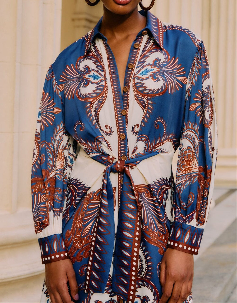 Someone stands before cream-colored columns wearing the Farm Rio Arabesque Scarf Blue Midi Dress, featuring a tied waist.