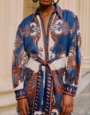 Someone stands before cream-colored columns wearing the Farm Rio Arabesque Scarf Blue Midi Dress, featuring a tied waist.