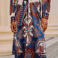 A person wears the Farm Rio Arabesque Scarf Blue Midi Dress, featuring a blue arabesque and paisley print with a tied waist, standing before cream-colored columns.