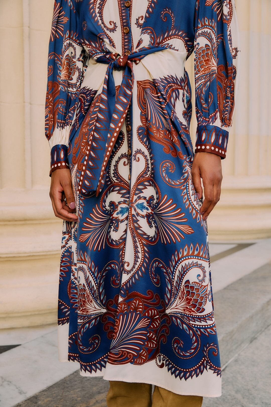 A person wears the Farm Rio Arabesque Scarf Blue Midi Dress, featuring a blue arabesque and paisley print with a tied waist, standing before cream-colored columns.