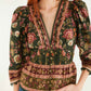 A woman stands against a plain background wearing the Farm Rio Flowering Garden Tree Green Blouse, featuring floral patterns, a deep V neckline, and puffed sleeves, paired with blue jeans.