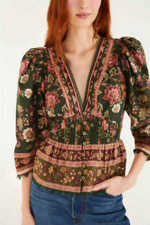 A woman stands against a plain background wearing the Farm Rio Flowering Garden Tree Green Blouse, featuring floral patterns, a deep V neckline, and puffed sleeves, paired with blue jeans.