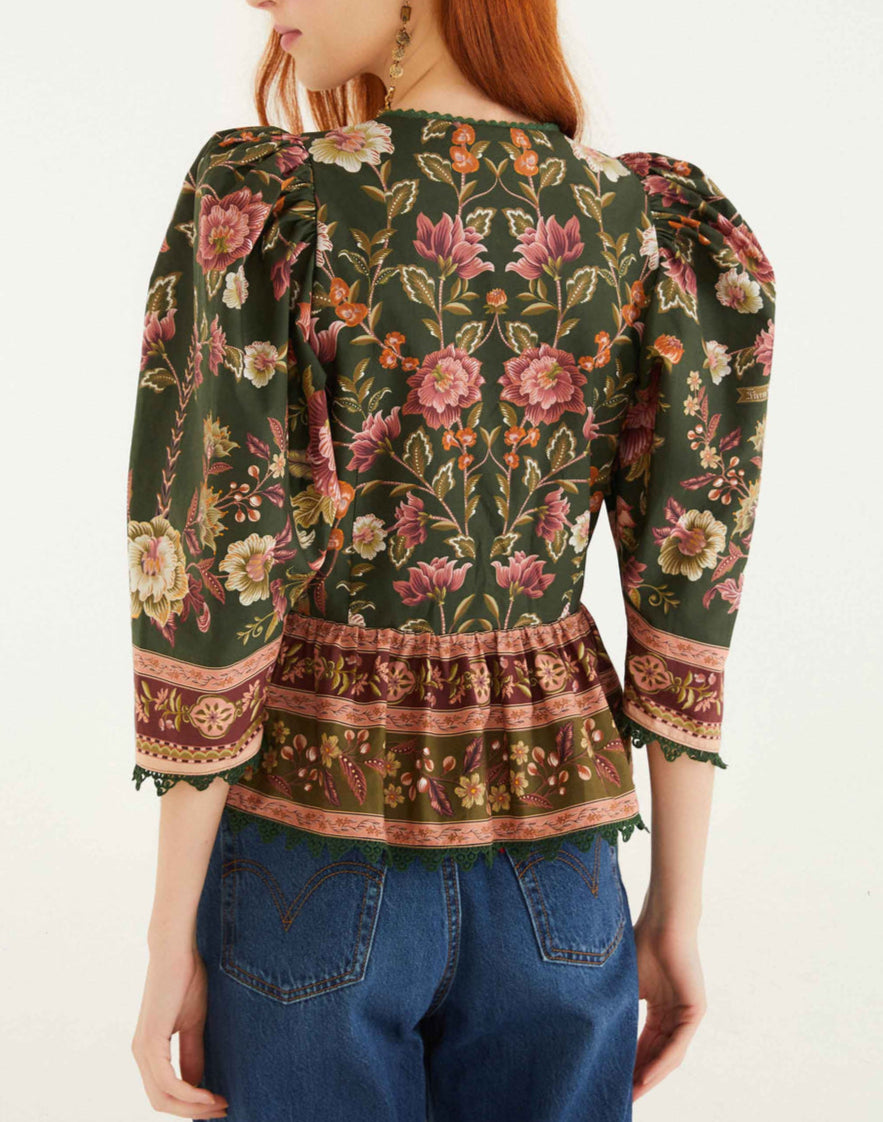 A woman with long red hair is seen from the back wearing the Farm Rio Flowering Garden Tree Green Blouse, which features floral patterns and puffed sleeves, paired with blue jeans.
