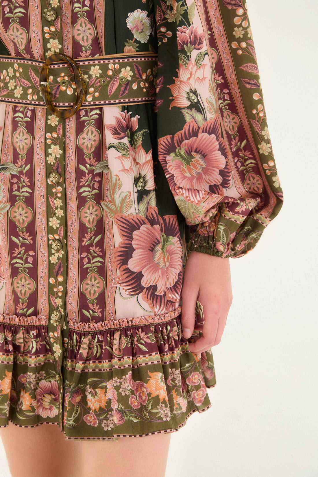 Close-up of the Flowering Garden Tree Green Mini Dress by Farm Rio, featuring floral and geometric patterns in pink, green, and brown, a belted waist with round buckle, and a ruffled hem.