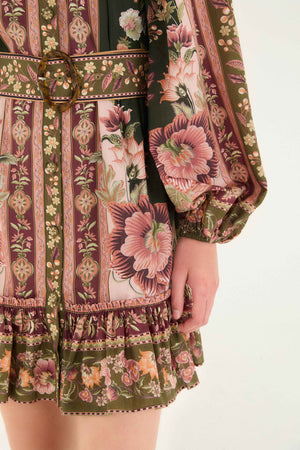 Close-up of the Flowering Garden Tree Green Mini Dress by Farm Rio, featuring floral and geometric patterns in pink, green, and brown, a belted waist with round buckle, and a ruffled hem.