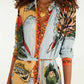 A woman wears the Farm Rio Beaches Path Multicolor Maxi Dress, adorned with vibrant map and botanical prints. She accessorizes with a chunky gold necklace and a shell pendant, her face partially out of frame to emphasize the dress’s striking tropical pattern.