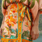 Person wearing a tropical-themed outfit with green, orange, and yellow palm tree and floral patterns, featuring the Farm Rio Birds Canto Dos Passardos Skirt, standing outdoors near water.