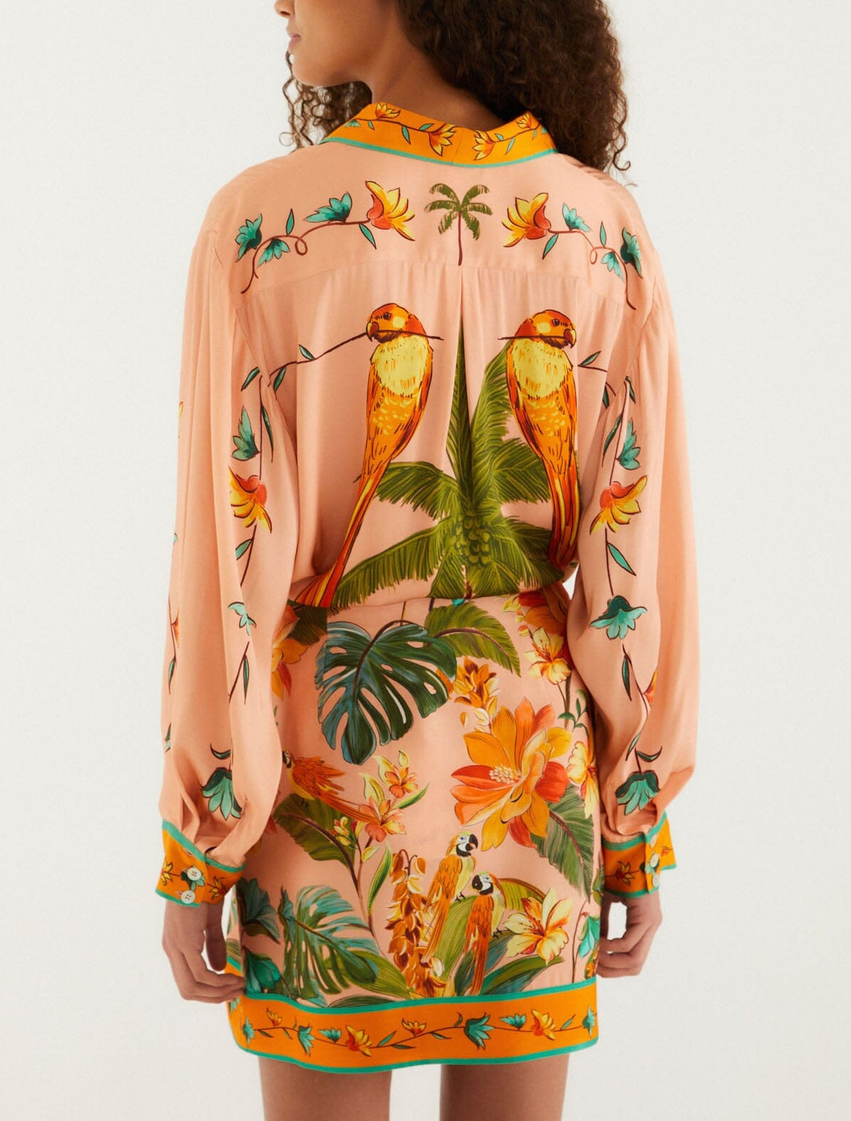 A woman faces away, wearing a peach dress and the FARM Rio Birds Canto Dos Passardos Skirt—a vibrant mini skirt with tropical yellow, green, and orange prints inspired by colorful birds.
