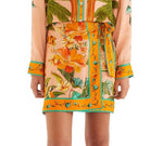 A person wears a peach floral dress with orange, yellow, and green tropical patterns, paired with the Farm Rio Birds Canto Dos Passardos Skirt and a matching tie belt, shown from shoulders to mid-thigh.