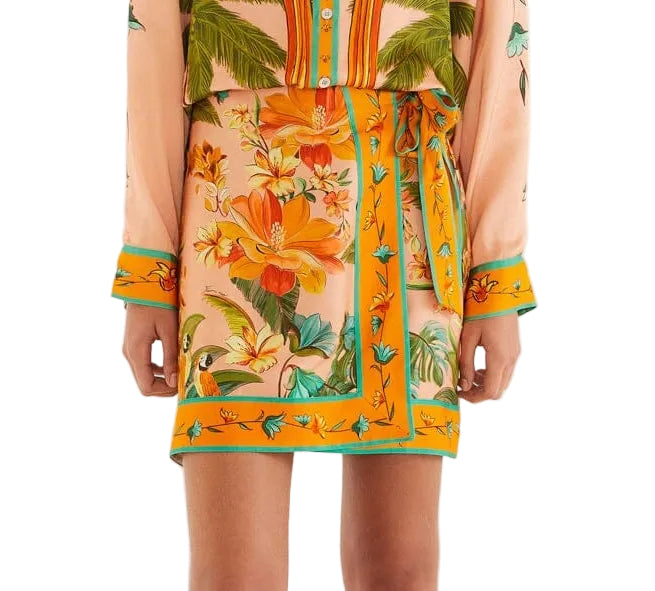 A person wears a peach floral dress with orange, yellow, and green tropical patterns, paired with the Farm Rio Birds Canto Dos Passardos Skirt and a matching tie belt, shown from shoulders to mid-thigh.