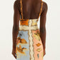 A person stands facing away, wearing the Farm Rio Rio Souvenir Mixed Scarf Multicolor Mini Dress, a sleeveless mini dress with rope straps and a pastel-toned print featuring shells, waves, and landscapes. The background is plain white.