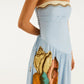 A woman models the Farm Rio Rio Souvenir Mixed Scarf Multicolor Maxi Dress, a strapless design featuring vibrant seashell and tropical prints with side lace-up detail, exuding resort elegance against a plain backdrop.