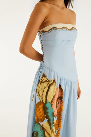 A woman models the Farm Rio Rio Souvenir Mixed Scarf Multicolor Maxi Dress, a strapless design featuring vibrant seashell and tropical prints with side lace-up detail, exuding resort elegance against a plain backdrop.