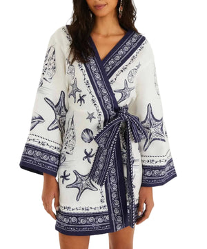 A woman models the Coast Scarf Off White Mini Dress by Farm Rio, featuring blue seashell and starfish patterns, wide sleeves, and a matching belt at the waist.
