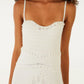A woman wearing the Farm Rio Palm Tree Knit Off White Midi Dress—a sleeveless, textured piece with thin straps and palm tree-inspired patterns—paired with large round earrings.