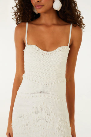 A woman wearing the Farm Rio Palm Tree Knit Off White Midi Dress—a sleeveless, textured piece with thin straps and palm tree-inspired patterns—paired with large round earrings.