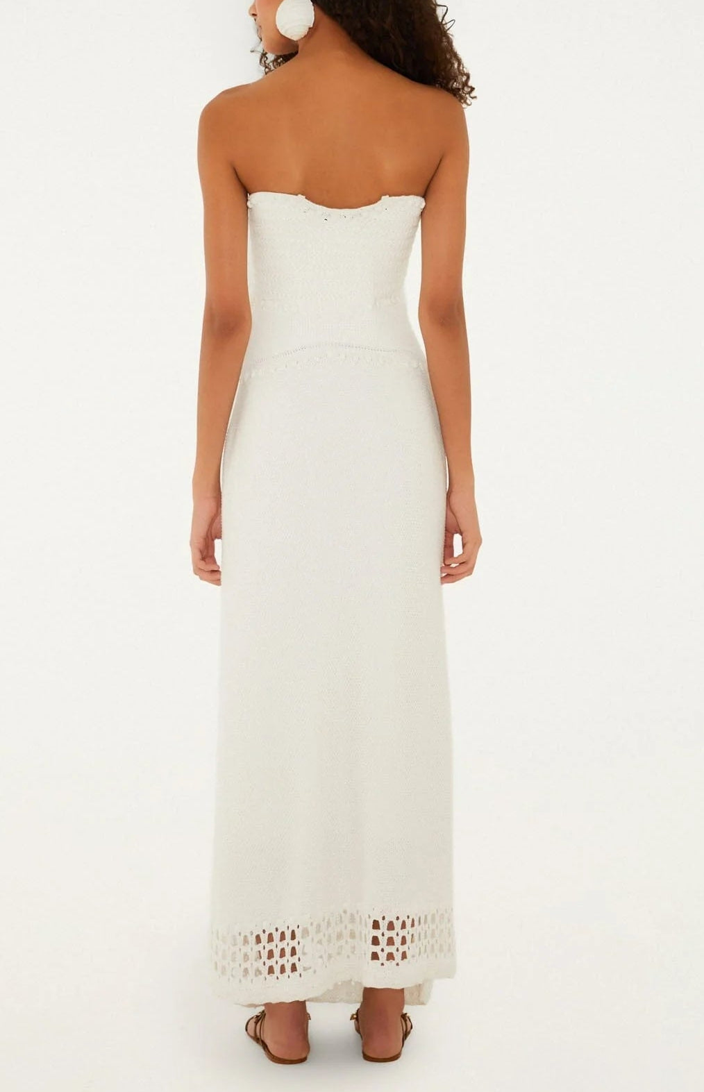 A woman facing away, wearing the Farm Rio Palm Tree Knit Off White Midi Dress with crochet hem detail, paired with sandals and large round earrings against a plain light background.
