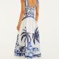 A woman is seen from the back wearing Farm Rio’s Wonderful City Mix Off White Maxi Dress featuring blue palm tree and geometric patterns. She completes the sleeveless look with sandals and earrings.