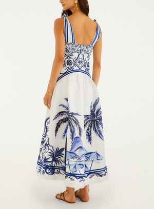A woman is seen from the back wearing Farm Rio’s Wonderful City Mix Off White Maxi Dress featuring blue palm tree and geometric patterns. She completes the sleeveless look with sandals and earrings.