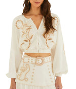 A woman wears the Richilieur Off White Blouse by Farm Rio, a long sleeve crop top with cut-out embroidery, paired with a matching high-waisted skirt featuring a wide belt and decorative stitching.