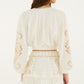 A woman stands against a plain background wearing the Farm Rio Richilieur Off White Blouse, featuring long sleeves and cut-out embroidery on the sleeves and waistband. Her brown hair is down and she faces away from the camera.