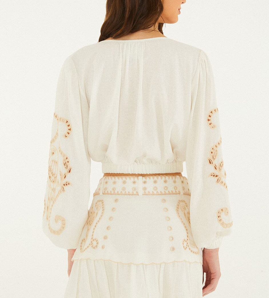 A woman stands against a plain background wearing the Farm Rio Richilieur Off White Blouse, featuring long sleeves and cut-out embroidery on the sleeves and waistband. Her brown hair is down and she faces away from the camera.