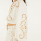 A woman wears the Farm Rio Richilieur Off White Blouse, a long-sleeve top with cut-out embroidery, paired with a matching skirt featuring decorative patterns and a wide belt.