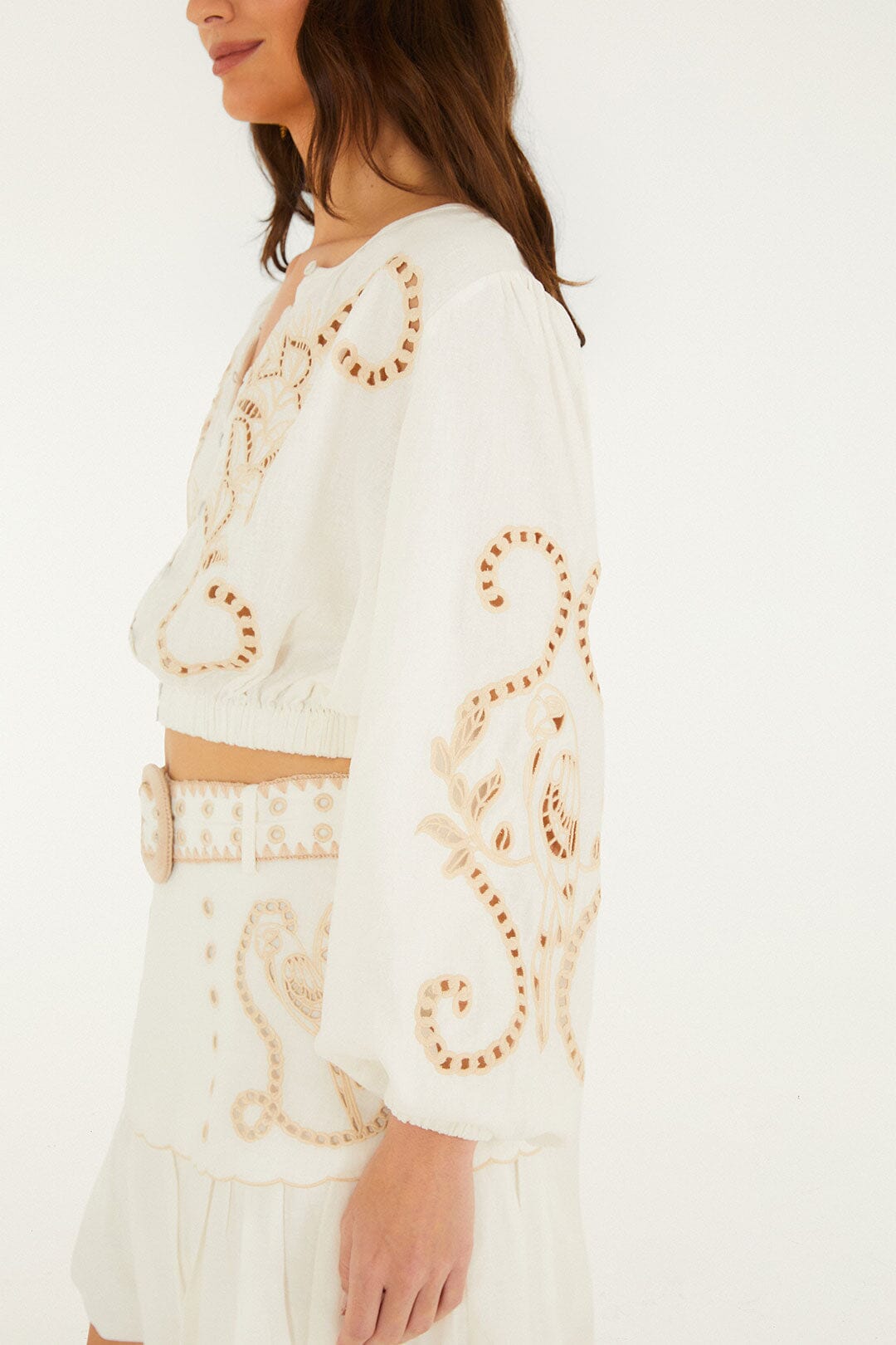 A woman wears the Farm Rio Richilieur Off White Blouse, a long-sleeve top with cut-out embroidery, paired with a matching skirt featuring decorative patterns and a wide belt.