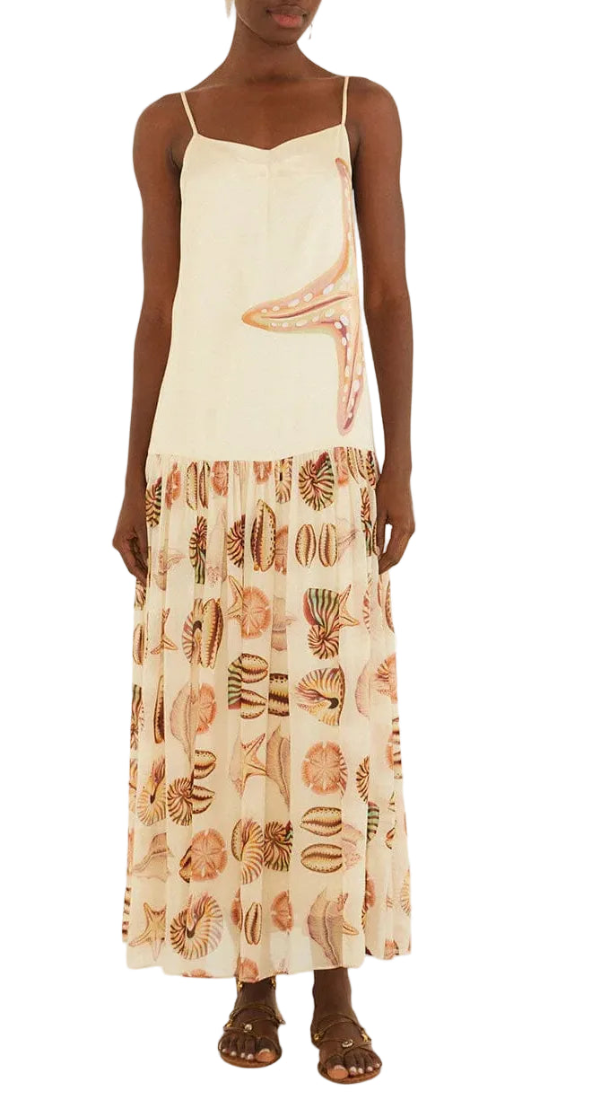A woman wearing the Farm Rio Marine Shells Sand Sleeveless Maxi Dress, featuring a sleeveless cream design with a starfish motif on the bodice and a flowing skirt adorned with a marine shell print for an effortless coastal look, styled with brown sandals.